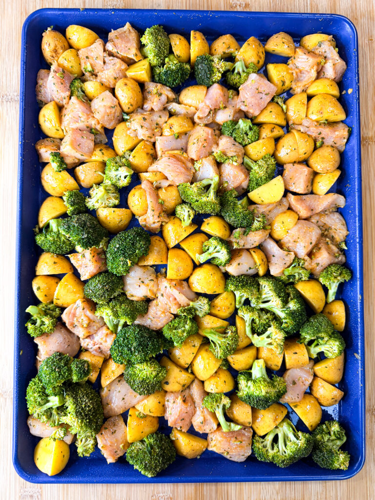 A blue baking sheet filled with seasoned raw chicken pieces, broccoli florets, and yellow potato chunks, all spread out evenly and ready for roasting.