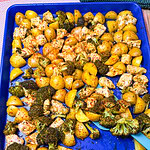 A blue baking tray filled with roasted broccoli, diced potatoes, and chicken, all seasoned with herbs and spices. A blue spatula rests in the bottom right corner.