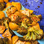 A blue spatula scoops up roasted broccoli, seasoned potato chunks, and pieces of chicken, all covered with spices, against a blue background.