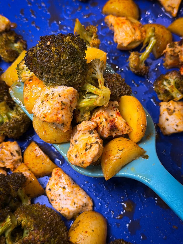A blue spatula holds roasted broccoli, potatoes, and seasoned chicken chunks, with more pieces scattered on a bright blue baking sheet.