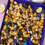A blue baking tray filled with roasted chicken, broccoli, and yellow potatoes, garnished with herbs and parmesan. A blue spoon rests on the tray, and lemon wedges are nearby.