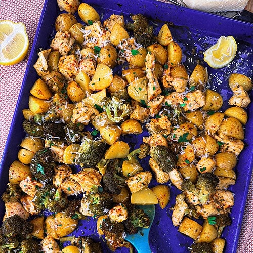 A blue baking tray filled with roasted chicken, broccoli, and yellow potatoes, garnished with herbs and parmesan. A blue spoon rests on the tray, and lemon wedges are nearby.