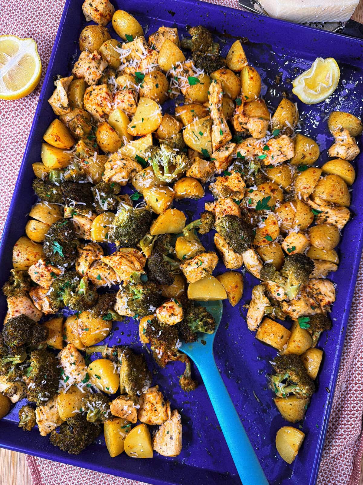 A blue baking tray filled with roasted chicken, broccoli, and yellow potatoes, garnished with herbs and parmesan. A blue spoon rests on the tray, and lemon wedges are nearby.