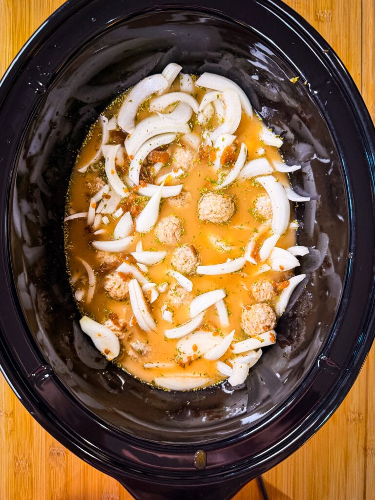 A slow cooker filled with meatballs, sliced onions, broth, and seasonings, ready to cook. The crockpot sits on a wooden surface.