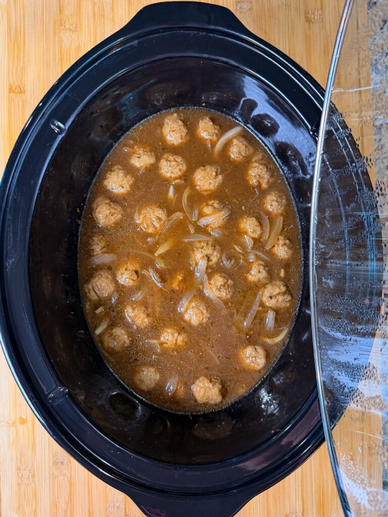 A slow cooker filled with meatballs, sliced onions, and brown broth, with the lid partially open, sits on a wooden surface.