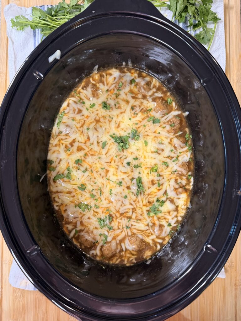 Slow cooker filled with melted cheese and garnished with chopped herbs on top of a savory brown dish, placed on a wooden surface with fresh parsley nearby.