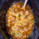 Meatballs in brown sauce with melted cheese and herbs, in a black slow cooker. A wooden spoon rests inside, partially mixing the ingredients. The mixture looks warm and freshly cooked.