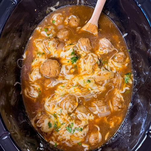Meatballs in brown sauce with melted cheese and herbs, in a black slow cooker. A wooden spoon rests inside, partially mixing the ingredients. The mixture looks warm and freshly cooked.