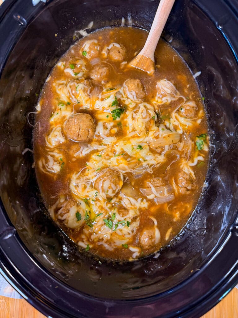 Meatballs in brown sauce with melted cheese and herbs, in a black slow cooker. A wooden spoon rests inside, partially mixing the ingredients. The mixture looks warm and freshly cooked.
