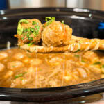 A wooden spoon holds two saucy meatballs with chopped herbs above a slow cooker filled with broth, noodles, and more meatballs.