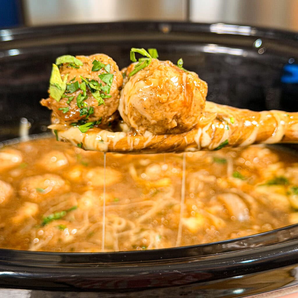 A ladle lifts two saucy meatballs topped with chopped herbs from a slow cooker filled with soup and noodles.