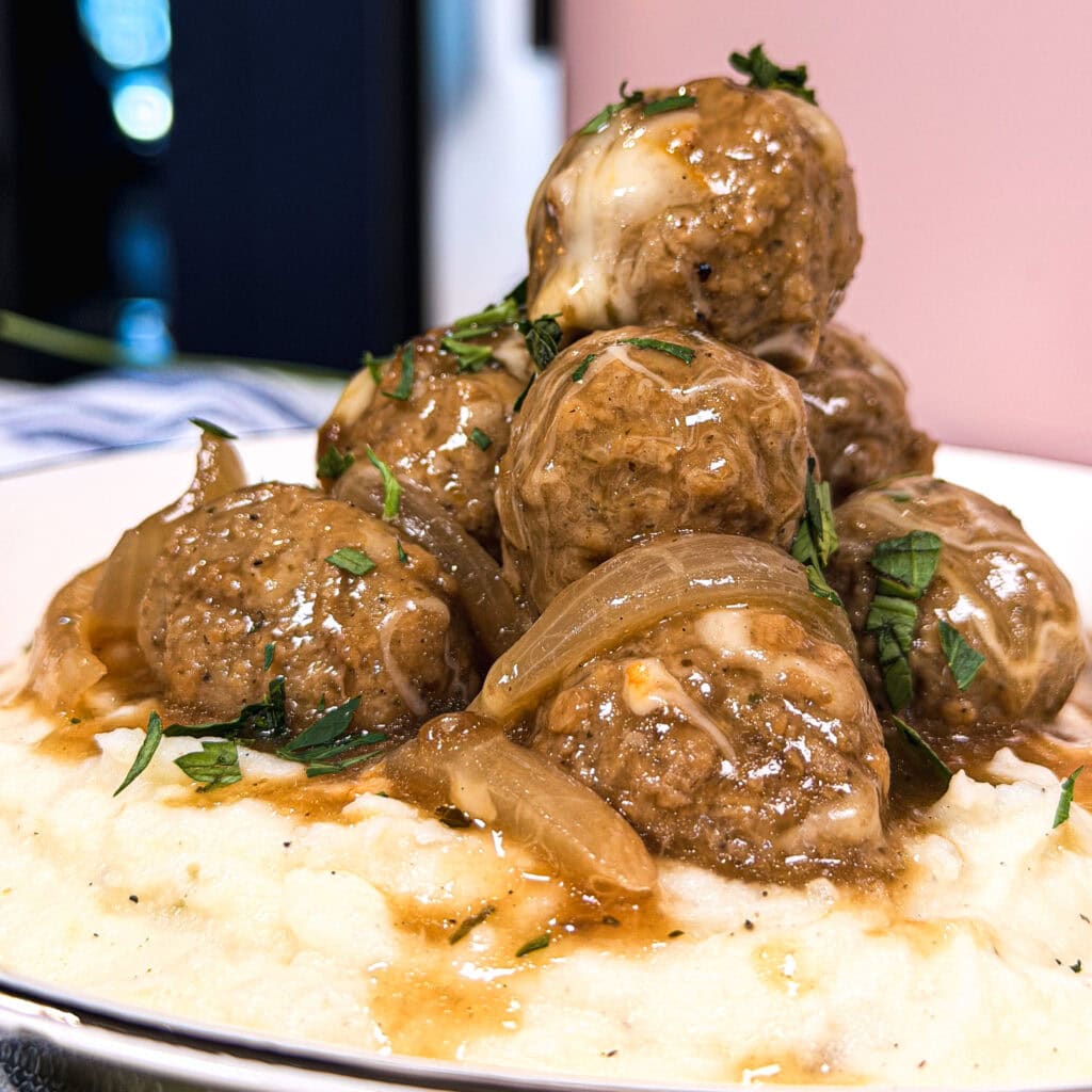 A plate of mashed potatoes topped with browned meatballs, sautéed onions, brown gravy, and garnished with chopped herbs.