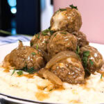 A plate of mashed potatoes topped with browned meatballs, sautéed onions, brown gravy, and garnished with chopped herbs.
