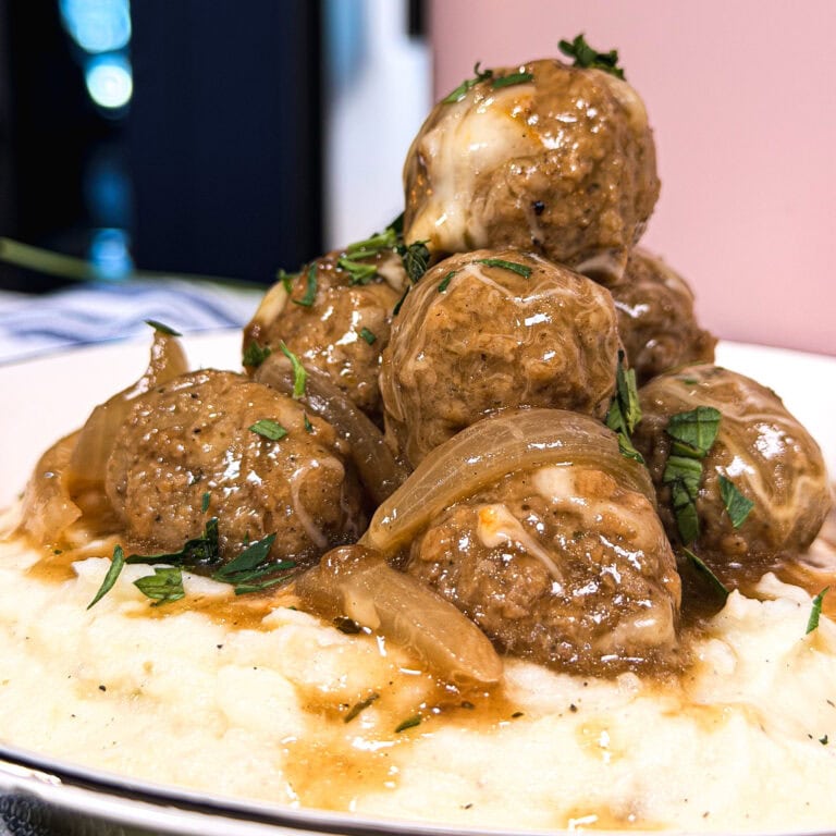 A plate of mashed potatoes topped with browned meatballs, sautéed onions, brown gravy, and garnished with chopped herbs.