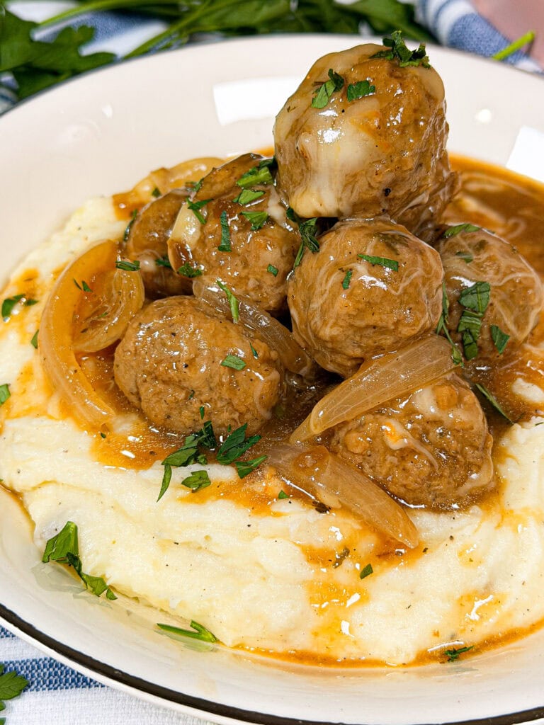 A bowl of creamy mashed potatoes topped with meatballs, sautéed onions, brown gravy, and sprinkled with chopped parsley.