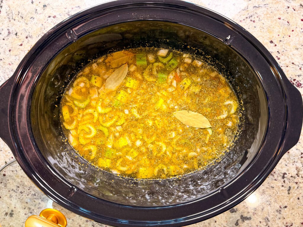A slow cooker filled with broth, chopped celery, onions, and spices with two bay leaves floating on top, sitting on a speckled countertop.