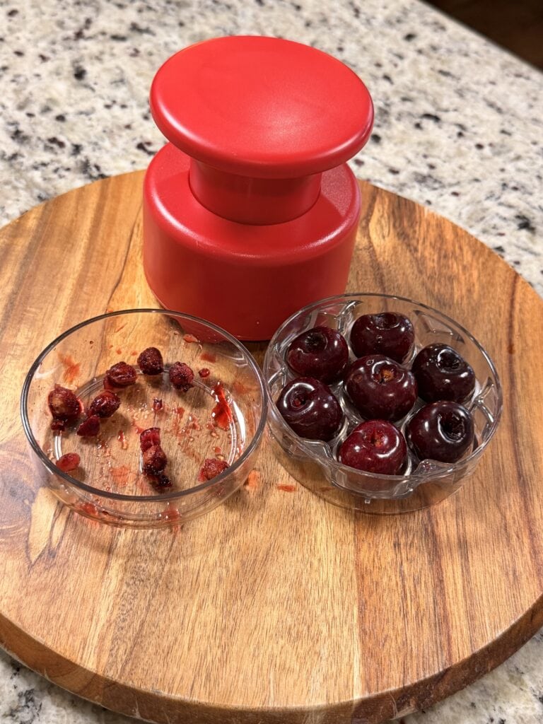 A red cherry pitter sits on a wooden board beside two glass bowls, one filled with pitted cherries and the other with cherry pits. The surface is granite.
