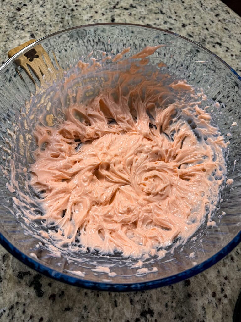 A glass bowl with swirled pink frosting sits on a granite countertop. A wooden spatula with some frosting left on it is resting at the edge of the bowl.