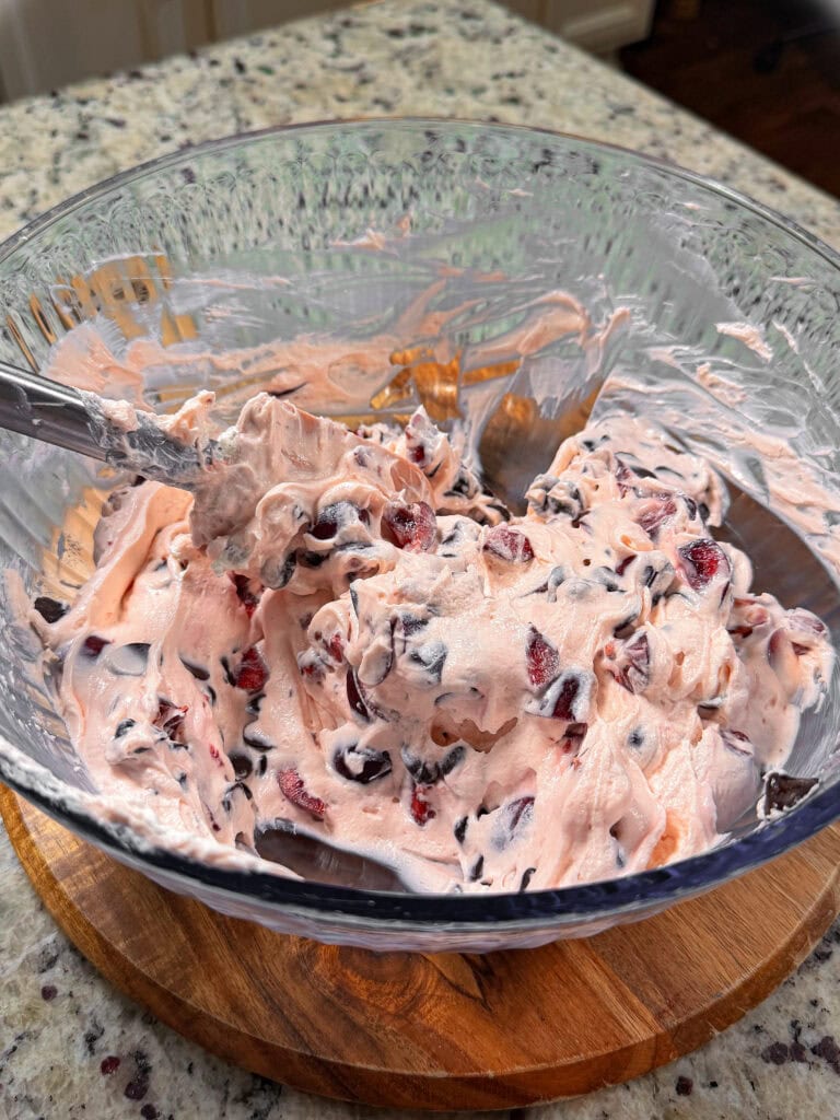 A glass bowl filled with a pink creamy mixture containing chopped cherries and chocolate chips, sitting on a wooden trivet on a granite countertop.