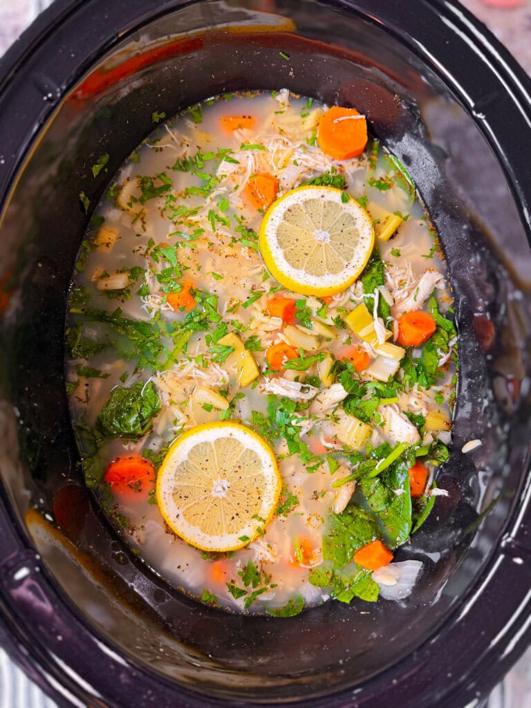 A slow cooker filled with chicken soup, showing sliced carrots, celery, spinach, herbs, shredded chicken, and two lemon slices floating on top in broth.