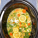 Chicken soup with sliced lemons, carrots, greens, and herbs in a black slow cooker, placed on a blue and white striped cloth.