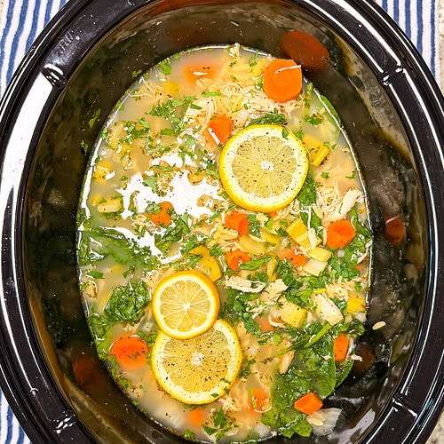 Chicken soup with sliced lemons, carrots, greens, and herbs in a black slow cooker, placed on a blue and white striped cloth.