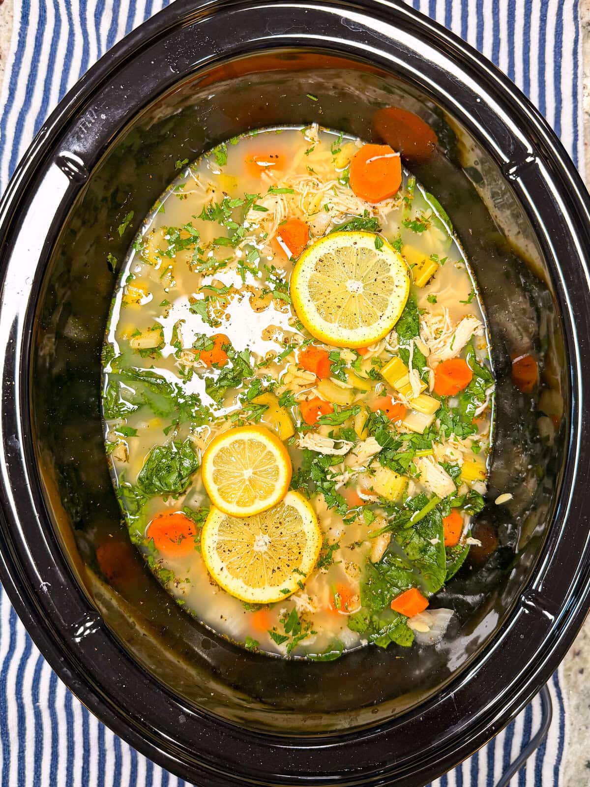 Chicken soup with sliced lemons, carrots, greens, and herbs in a black slow cooker, placed on a blue and white striped cloth.