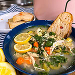 A blue bowl of chicken soup with lemon slices, carrots, celery, herbs, and shredded chicken, served with toasted bread. Behind it, a plate holds cornbread and a pink pot is partially visible.