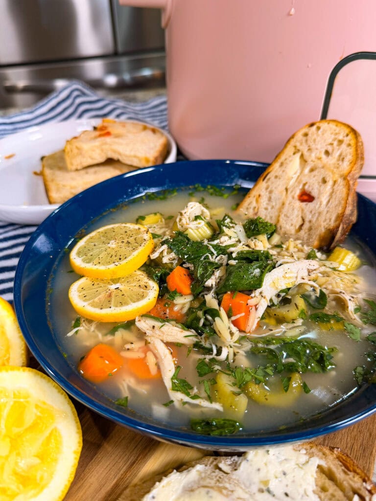 A blue bowl of chicken soup with lemon slices, carrots, celery, herbs, and shredded chicken, served with toasted bread. Behind it, a plate holds cornbread and a pink pot is partially visible.