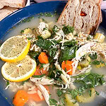 A bowl of chicken soup with shredded chicken, carrots, spinach, orzo pasta, lemon slices, and chopped herbs, served with a slice of bread.
