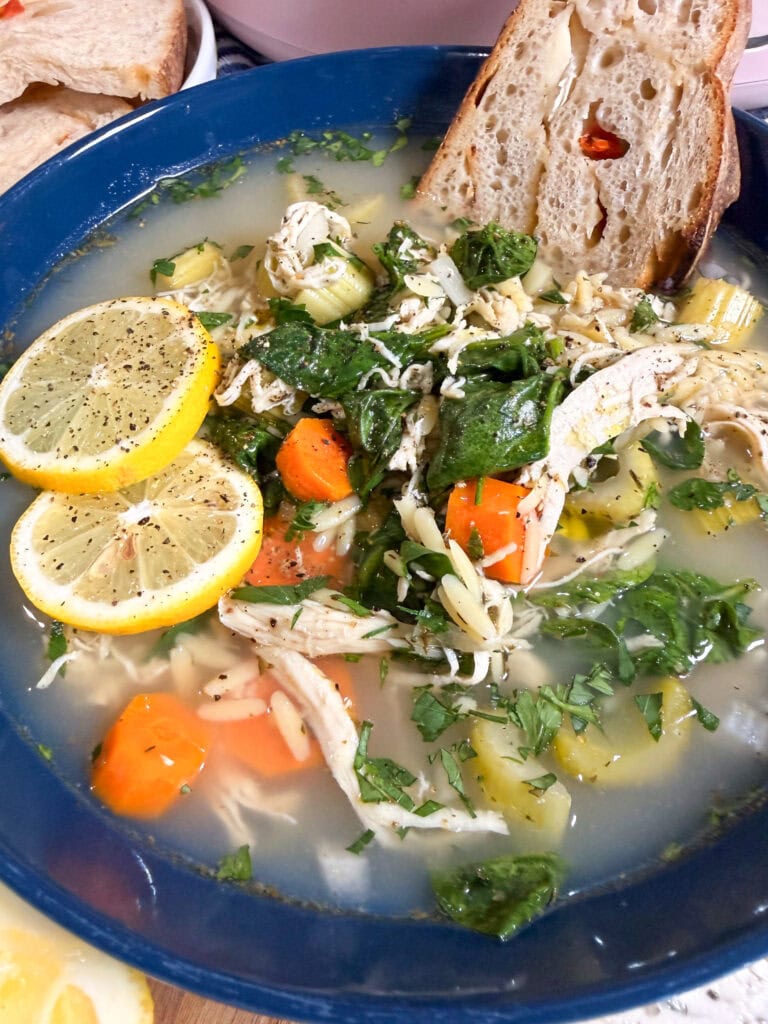 A bowl of chicken soup with shredded chicken, carrots, spinach, orzo pasta, lemon slices, and chopped herbs, served with a slice of bread.