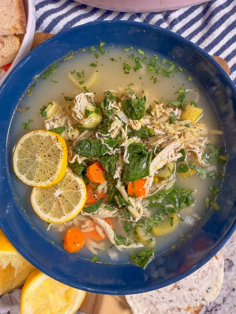 A blue bowl filled with chicken noodle soup, featuring shredded chicken, carrots, celery, spinach, herbs, and two lemon slices. The bowl is set on a striped cloth with bread and lemon wedges nearby.