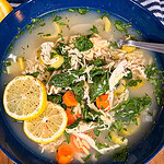 A bowl of chicken soup with carrots, spinach, celery, and shredded chicken, topped with two lemon slices and fresh herbs. Theres a spoon in the bowl and buttered bread on the side.