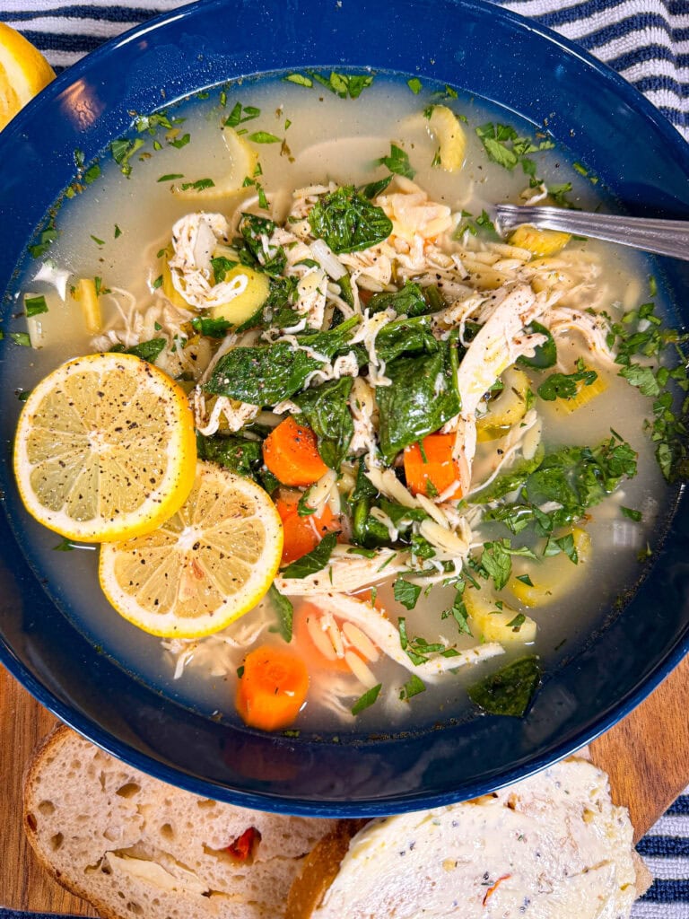 A bowl of chicken soup with carrots, spinach, celery, and shredded chicken, topped with two lemon slices and fresh herbs. Theres a spoon in the bowl and buttered bread on the side.