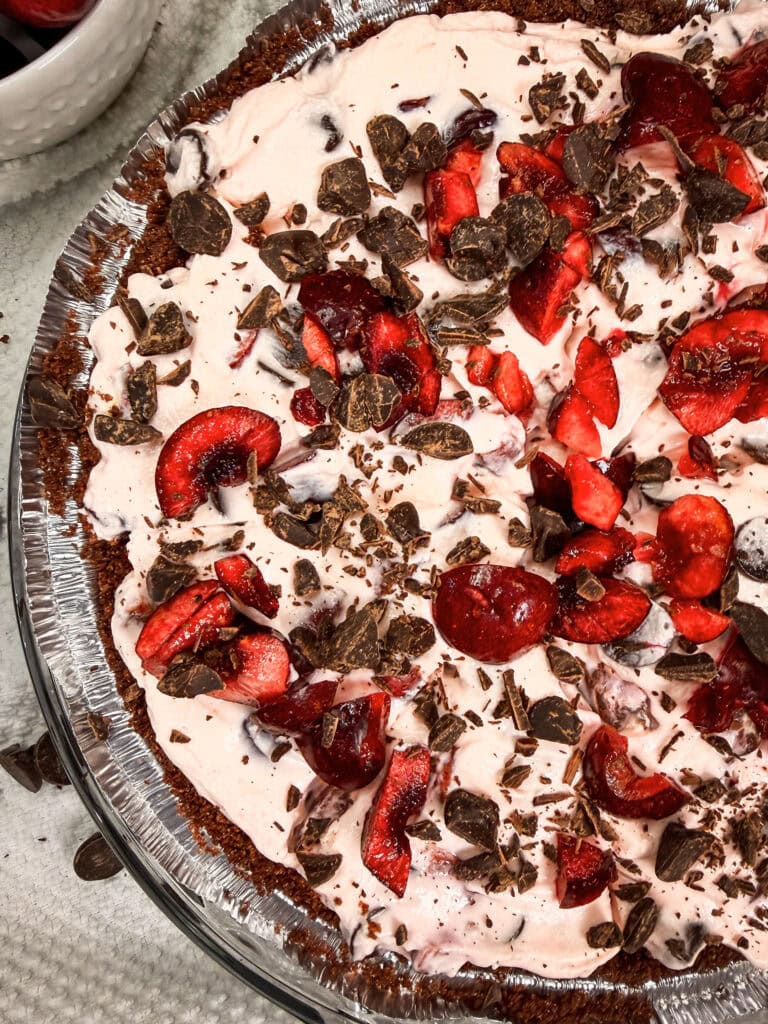 A close-up of a pie topped with whipped cream, fresh cherry slices, and chocolate shavings in a foil pie pan.