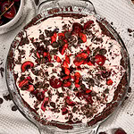 A glass pie dish filled with a chocolate crust, creamy pink filling, topped with fresh cherries, chocolate shavings, and chocolate chips. Whole and cut cherries and chips are scattered on a white cloth around the pie.