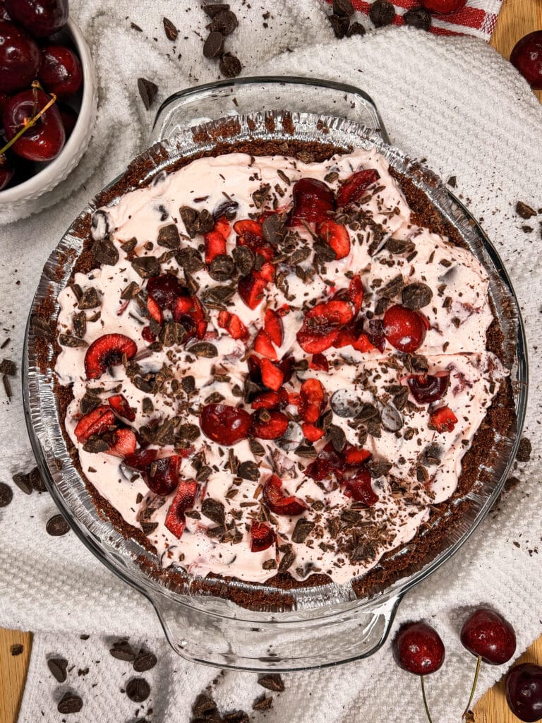 A glass pie dish filled with a chocolate crust, creamy pink filling, topped with fresh cherries, chocolate shavings, and chocolate chips. Whole and cut cherries and chips are scattered on a white cloth around the pie.