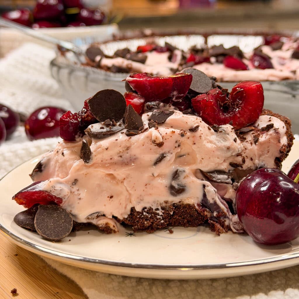 A slice of cherry chocolate chip pie with a chocolate crust, topped with fresh cherries and chocolate pieces, sits on a plate beside whole cherries.