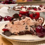 A slice of cherry chocolate chip pie with a chocolate crust, topped with fresh cherries and chocolate pieces, sits on a plate beside whole cherries.