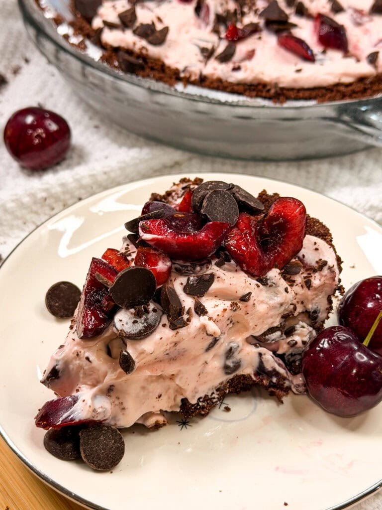 A slice of cherry chocolate cream pie on a plate, topped with fresh cherries and chocolate chips, with whole cherries on the side. The rest of the pie is visible in the background.