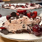 A slice of cherry chocolate chip ice cream pie topped with fresh cherries and chocolate chunks sits on a plate beside whole cherries.