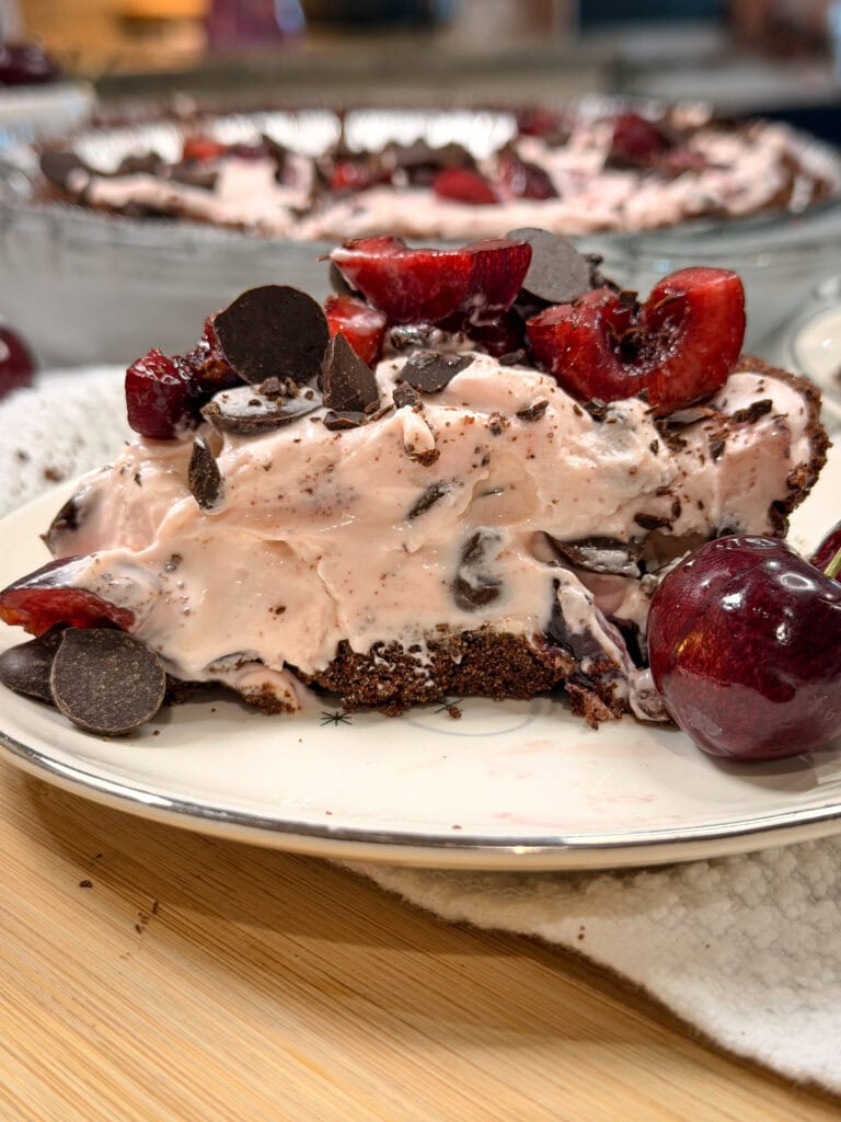 A slice of cherry chocolate chip ice cream pie topped with fresh cherries and chocolate chunks sits on a plate beside whole cherries.