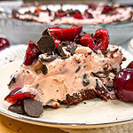 A slice of chocolate cherry cream pie topped with cherries and chocolate chips sits on a plate, with another whole pie and more cherries in the background.