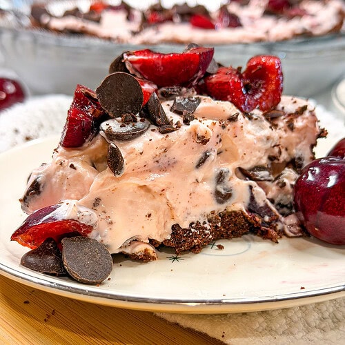 A slice of chocolate cherry cream pie topped with cherries and chocolate chips sits on a plate, with another whole pie and more cherries in the background.