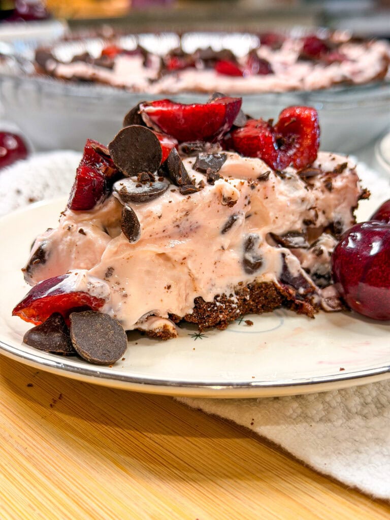 A slice of chocolate cherry cream pie topped with cherries and chocolate chips sits on a plate, with another whole pie and more cherries in the background.