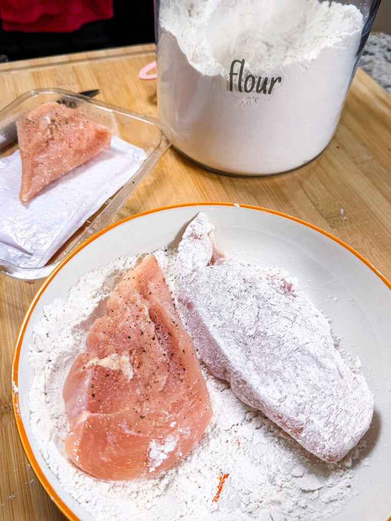 Raw chicken breasts on a plate, one covered in flour, the other being coated. A flour container and a package with more chicken are on a wooden surface in the background.