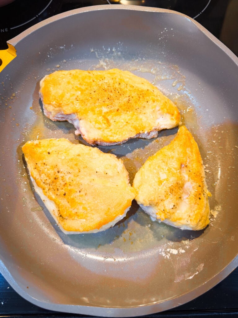 Three pieces of seasoned chicken breast are being cooked in a nonstick frying pan, with a golden-brown crust forming on the surface.