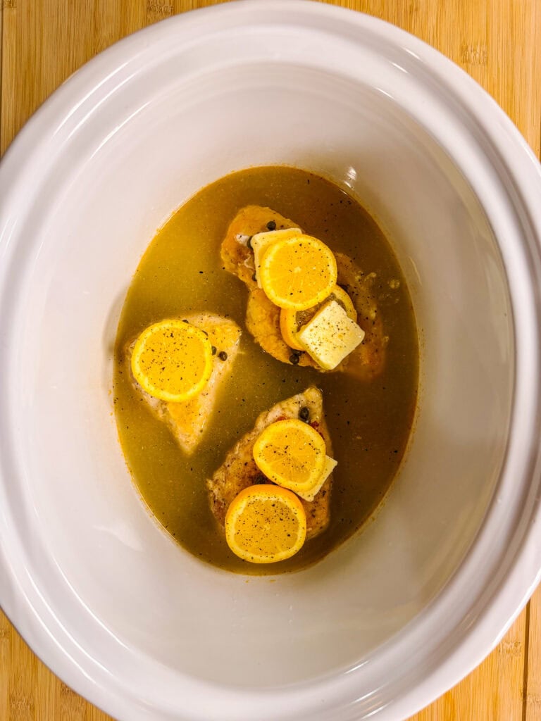 Raw chicken breasts topped with lemon slices, black pepper, and a pat of butter sit in broth inside a white slow cooker on a wooden surface.