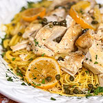 A plate of pasta topped with sliced chicken breast, lemon slices, capers, and chopped parsley, served on a white dish.