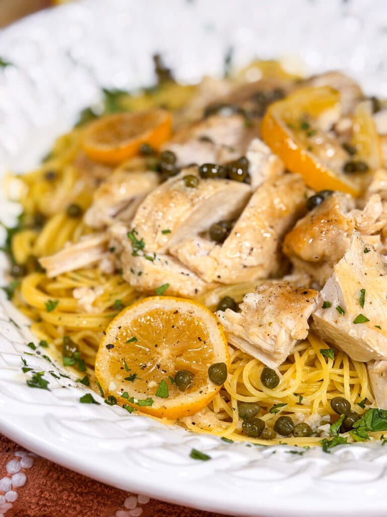 A plate of pasta topped with sliced chicken breast, lemon slices, capers, and chopped parsley, served on a white dish.
